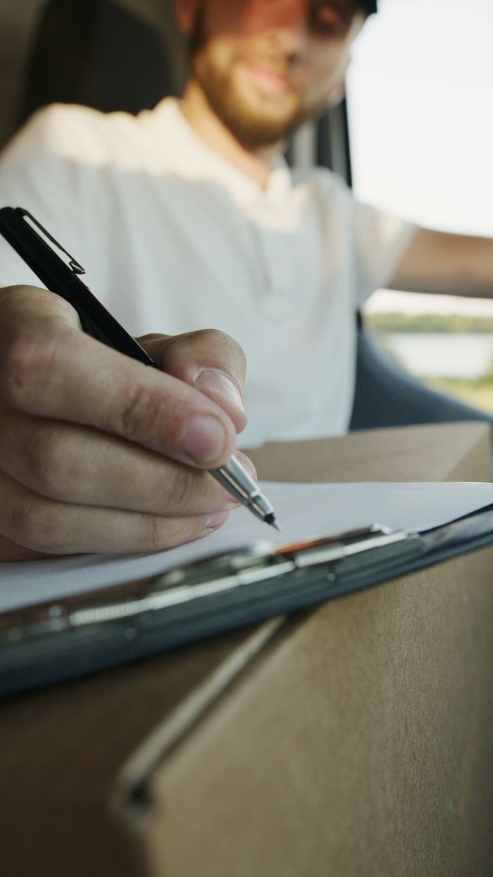 Mastering the First Impression: Your intriguing post title goes here Close-up of a delivery driver writing on a clipboard while holding a package inside a vehicle.